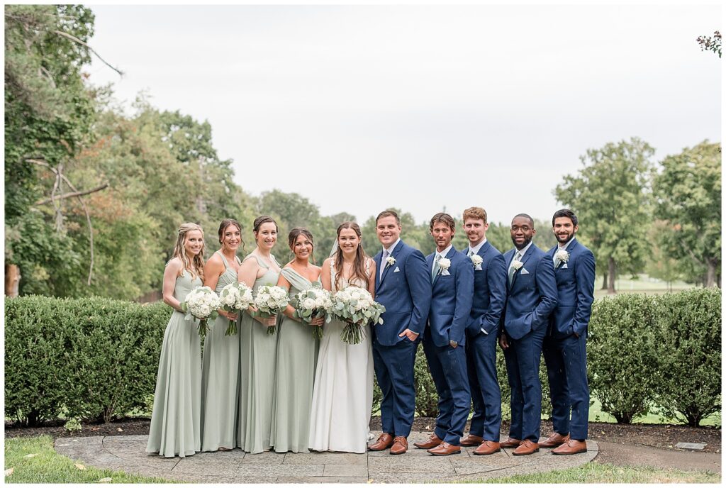 couple surrounded by their bridal party wearing sage green gowns and navy blue suits in flourtown pa