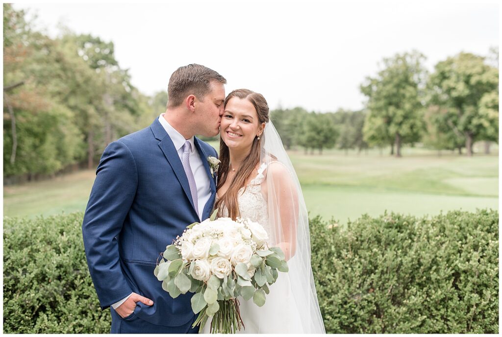 groom kissing his bride on her right cheek with flourtown golf club course behind them