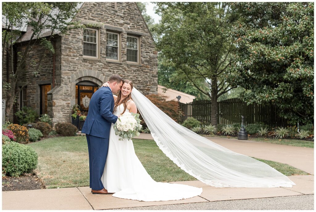 couple hugging by stone building with bride's long veil trailing behind her in flourtown pennsylvania