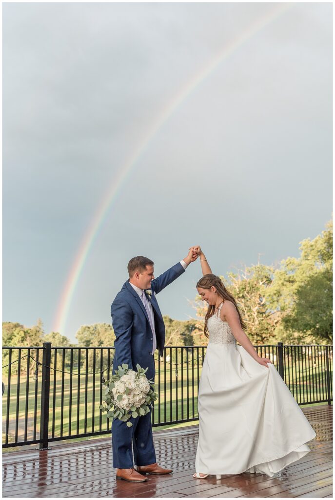 groom twirling his bride with rainbow in sky behind them at flourtown country club in pennsylvania