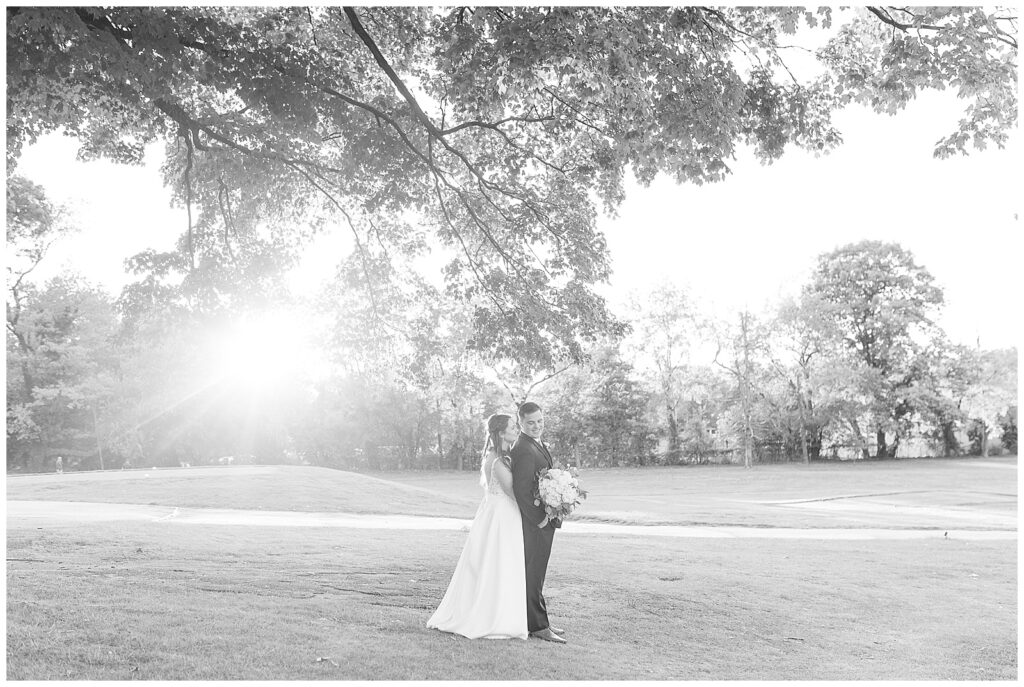 black and white photo of sun peeking through clouds as bride hugs groom from behind in pennsylvania