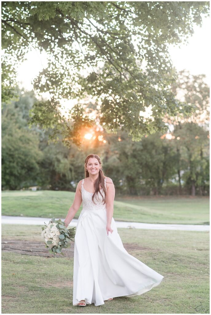 bride holding bouquet and walking towards camera with sun peeking through trees behind her in flourtown pennsylvania