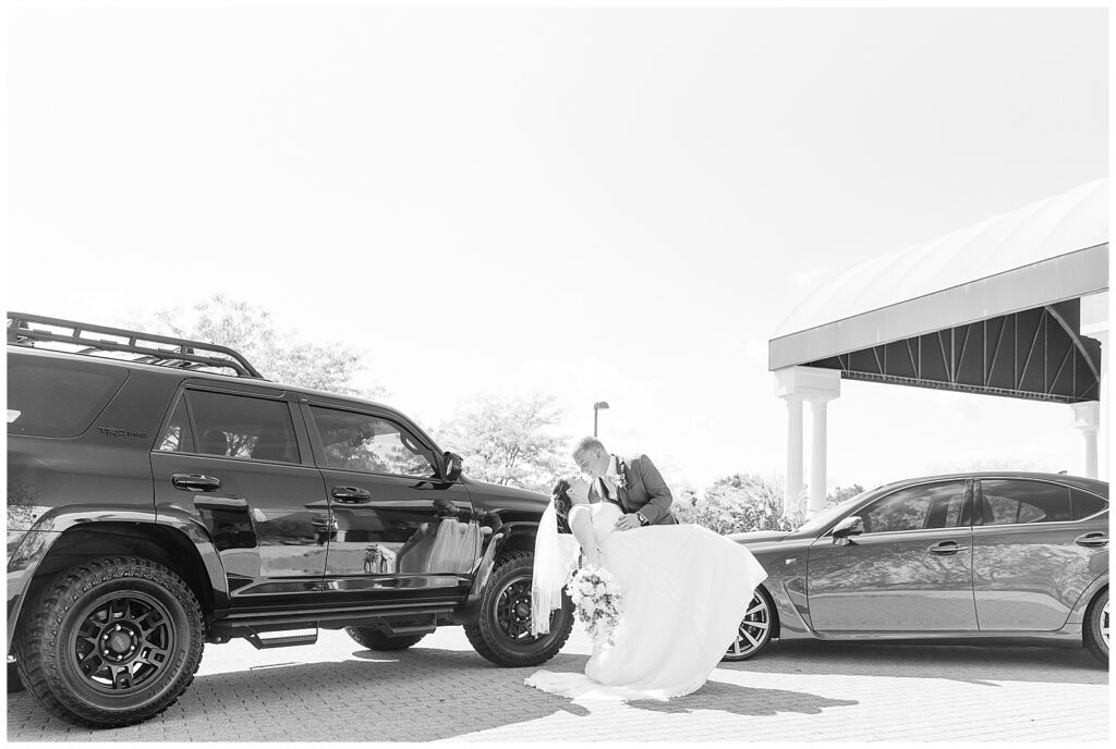 groom dipping his bride back as they kiss by two cars at jumping brook country club