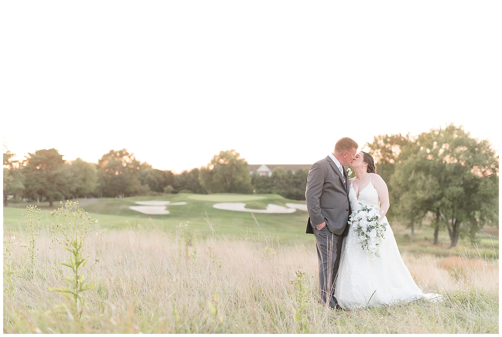 couple kissing standing in tall grasses with golf green behind them at jumping brook country club