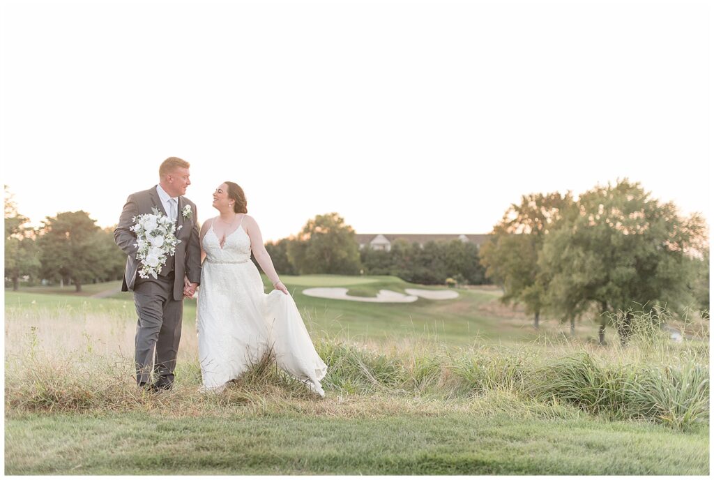 couple walking towards camera as bride holds dress train with golf course around them in new jersey