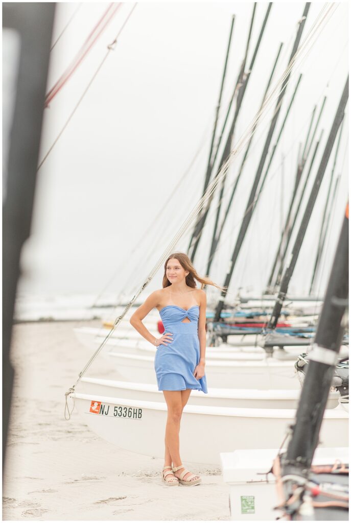 senior girl in blue dress with right hand on hip standing on beach by sailboat in new jersey