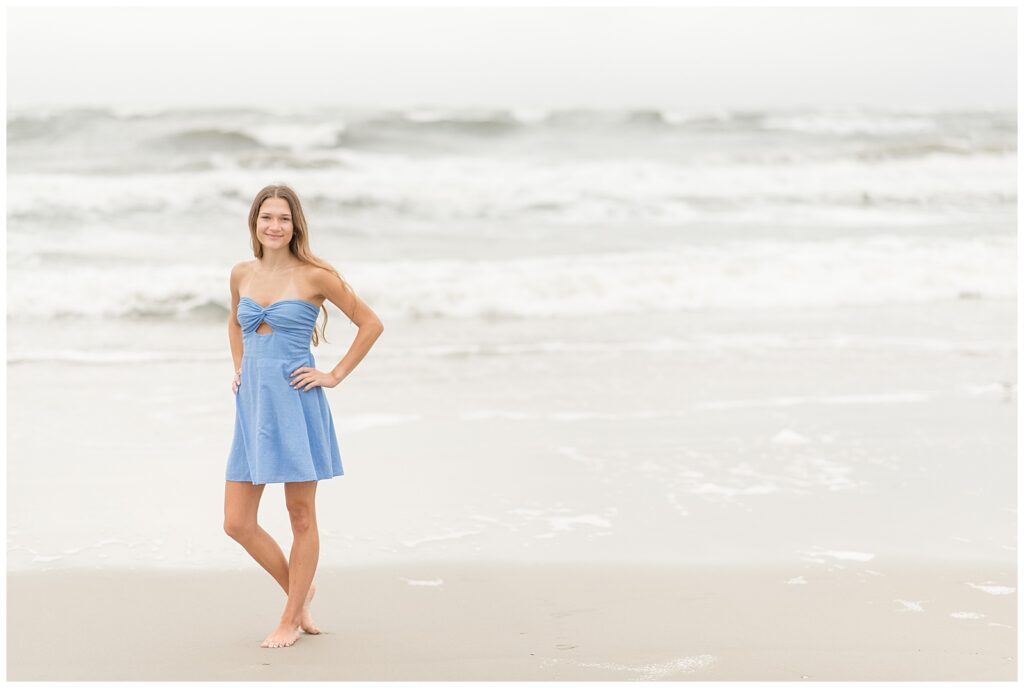 senior girl with hands on hips standing along the ocean on new jersey beach on overcast evening