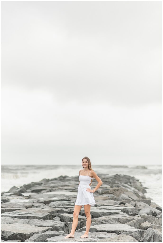 senior girl in white cress standing on large rocks that extend out into the atlantic ocean in new jersey