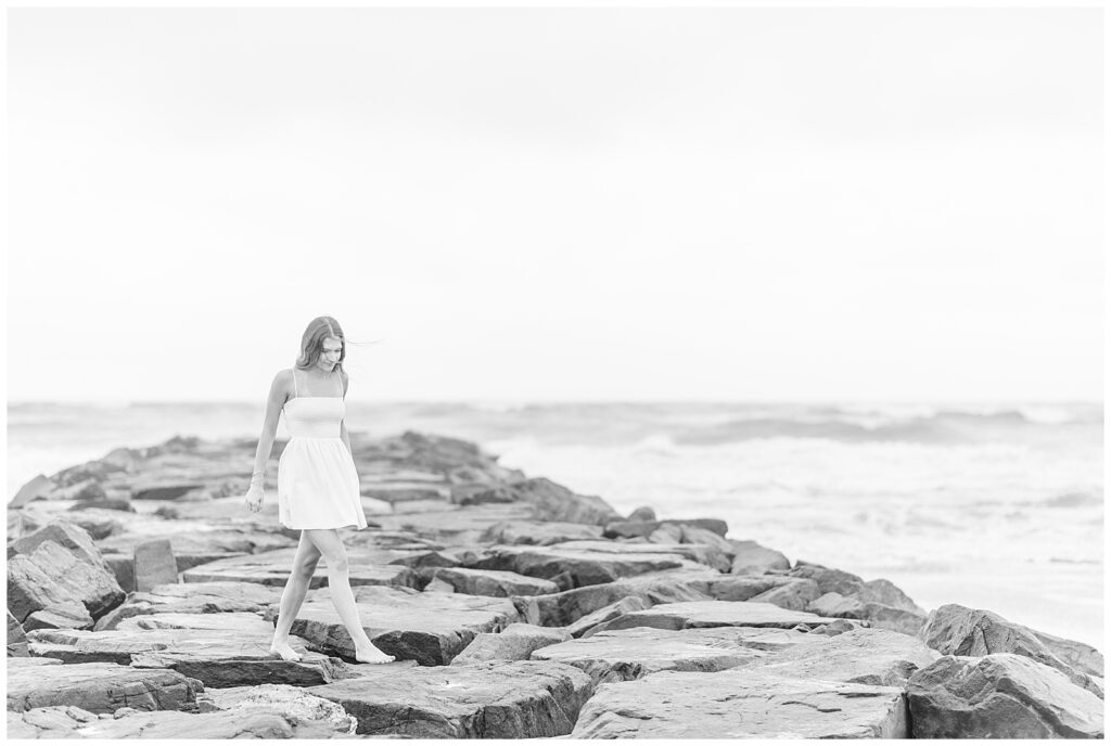 black and white photo of senior girl walking along large rocks at beach in new jersey