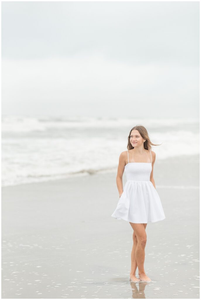 senior girl with hands in her white dress pockets and looking into distance by ocean in new jersey