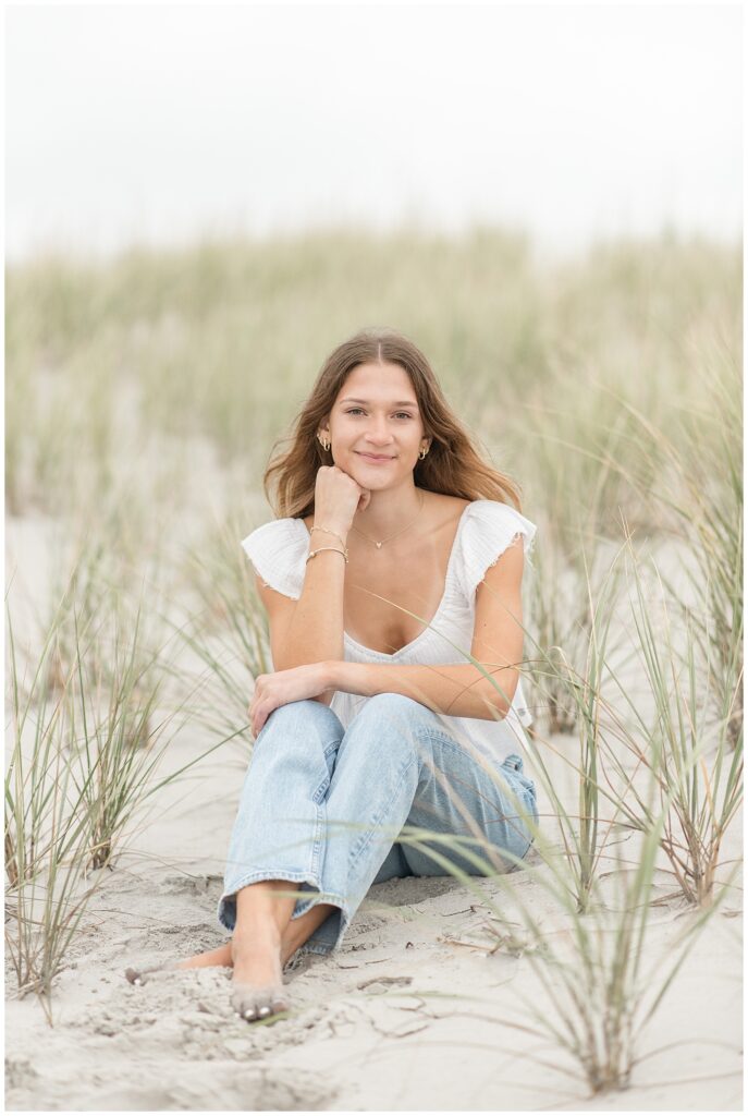 senior girl sitting in dunes with her right elbow on her knee as she rest her chin on her fist in new jersey