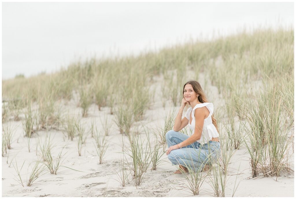 senior girl crouched down in dunes on overcast evening in new jersey