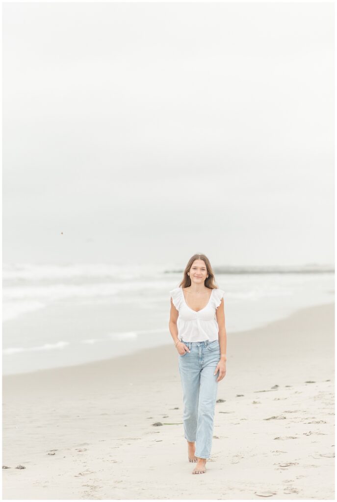 senior girl walking along beach in jeans and white tank top in new jersey