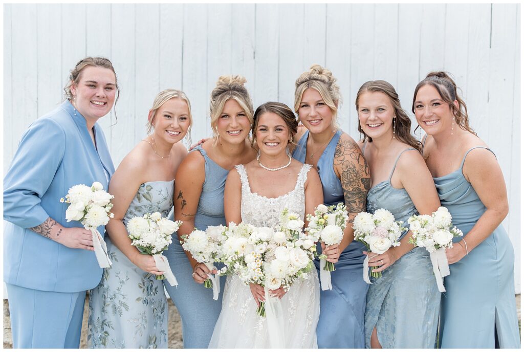 bride surrounded by six bridesmaids in light blue gowns all holding bouquets by white barn