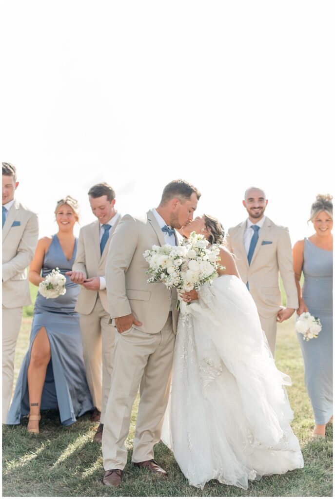 couple kissing with their wedding party behind them on sunny fall day at family farm in pennsylvania