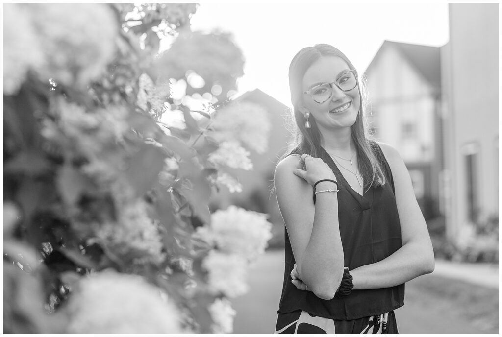 black and white photo of senior girl with right hand resting on right shoulder by flowering bush at stoudtburg village