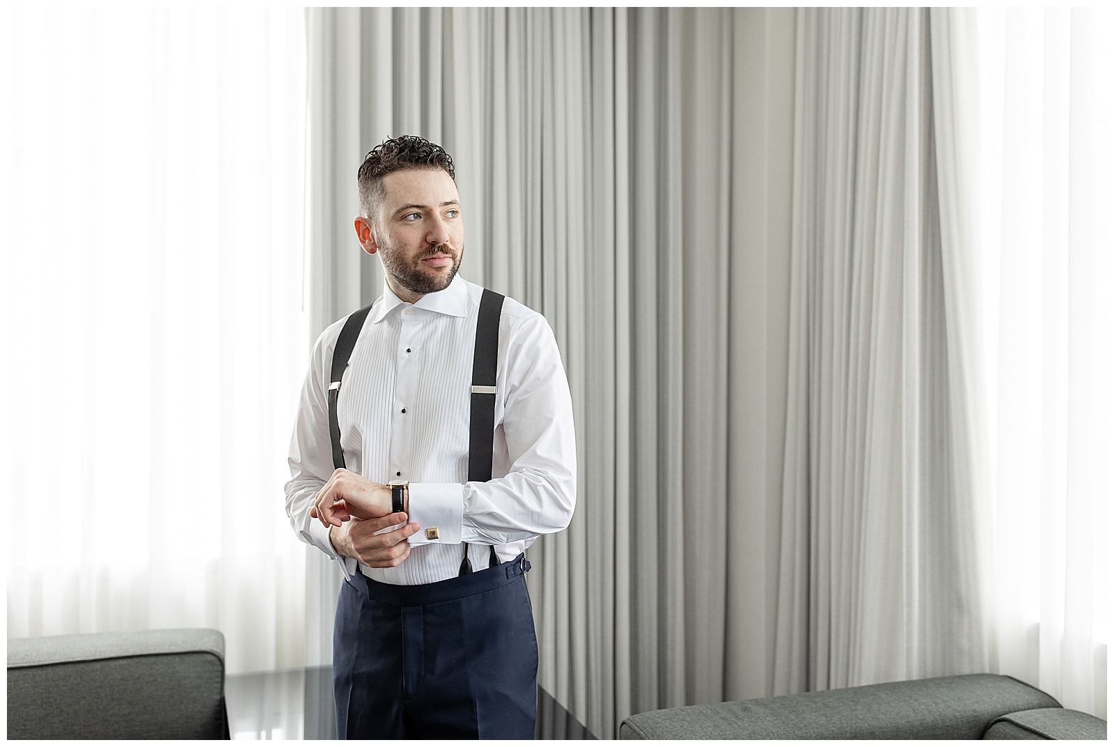 groom adjusting his wristwatch as he looks left in suit pants with white dress shirt and suspenders in philadelphia hotel room