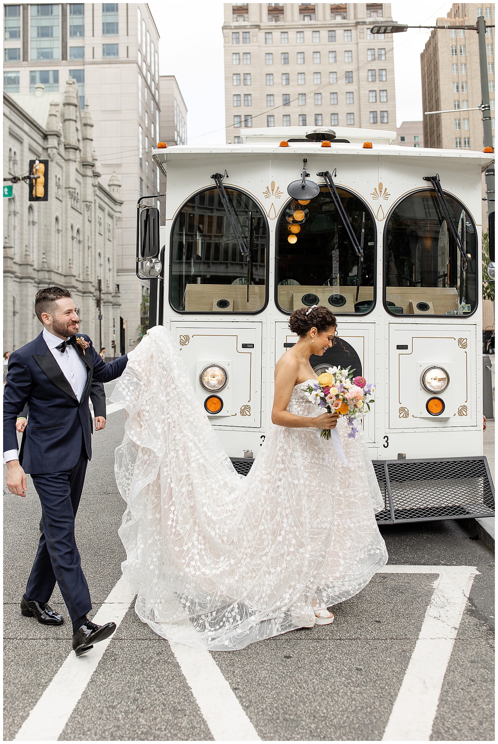 groom holding up bride's long dress train as they pass in front of white trolley on street in philadelphia