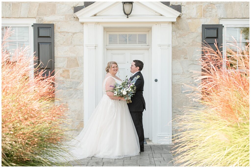 couple hugging as bride looks at camera in sleeveless white gown by white front door of stone house in lancaster county