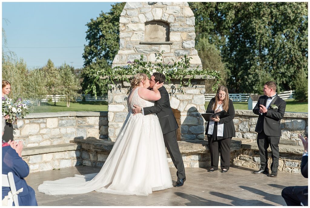 couple sharing their first kiss by stone fireplace during outdoor wedding ceremony on sunny fall day in lancaster pennsylvania