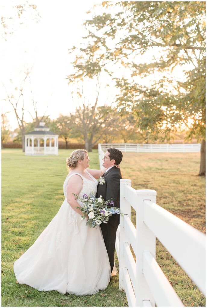 couple hugging as groom leans back against white fence at sunset with white gazebo in background in lancaster county