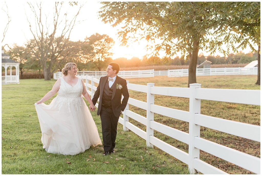 bride and groom holding hands and walking along white fence at the barn at silverstone at sunset