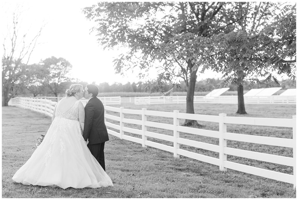 black and white photo of couple kissing with their backs toward the camera by white fence at the barn at silverstone