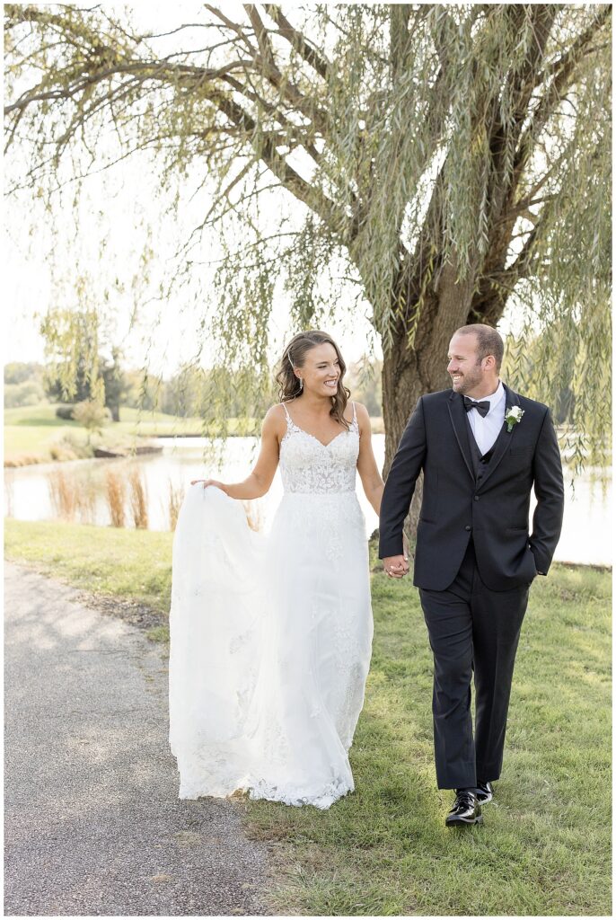 bride and groom holding hands as bride holds her long white dress train by willow tree at the links at gettysburg