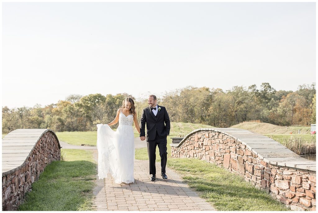 couple holding hands and bride holds dress train as they walk toward camera on golf course brick pathway at the links at gettysburg