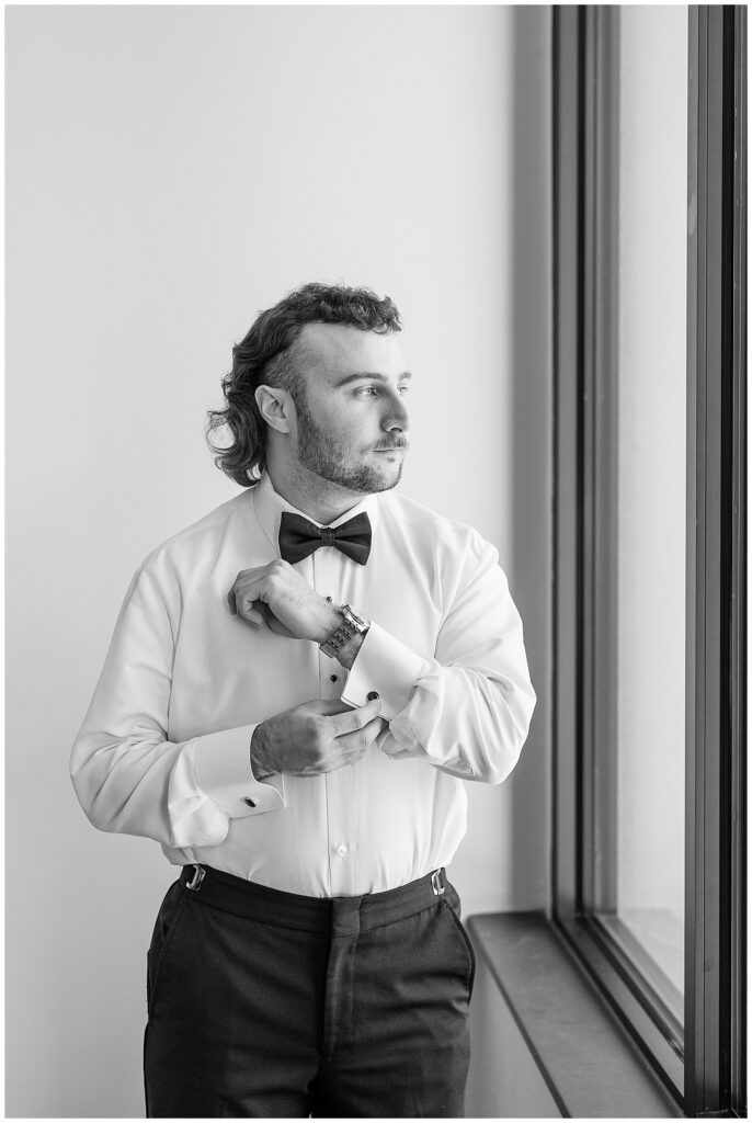 groom looking left out window and adjusting his cuff links in black and white photo on wedding day in york pennsylvania