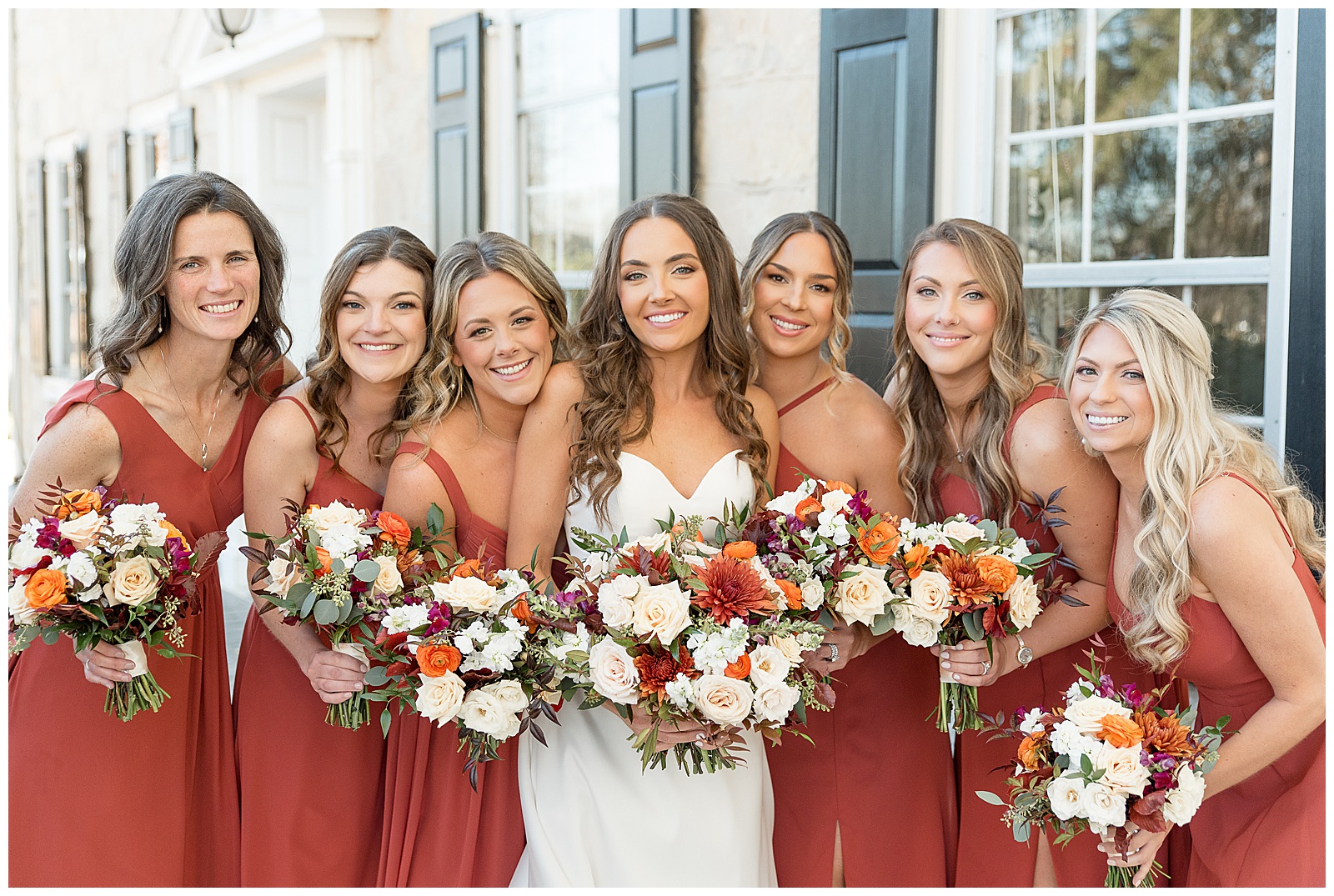 bride surrounded by her bridesmaids all wearing rust colored gowns and holding flower bouquets at the barn at silverstone