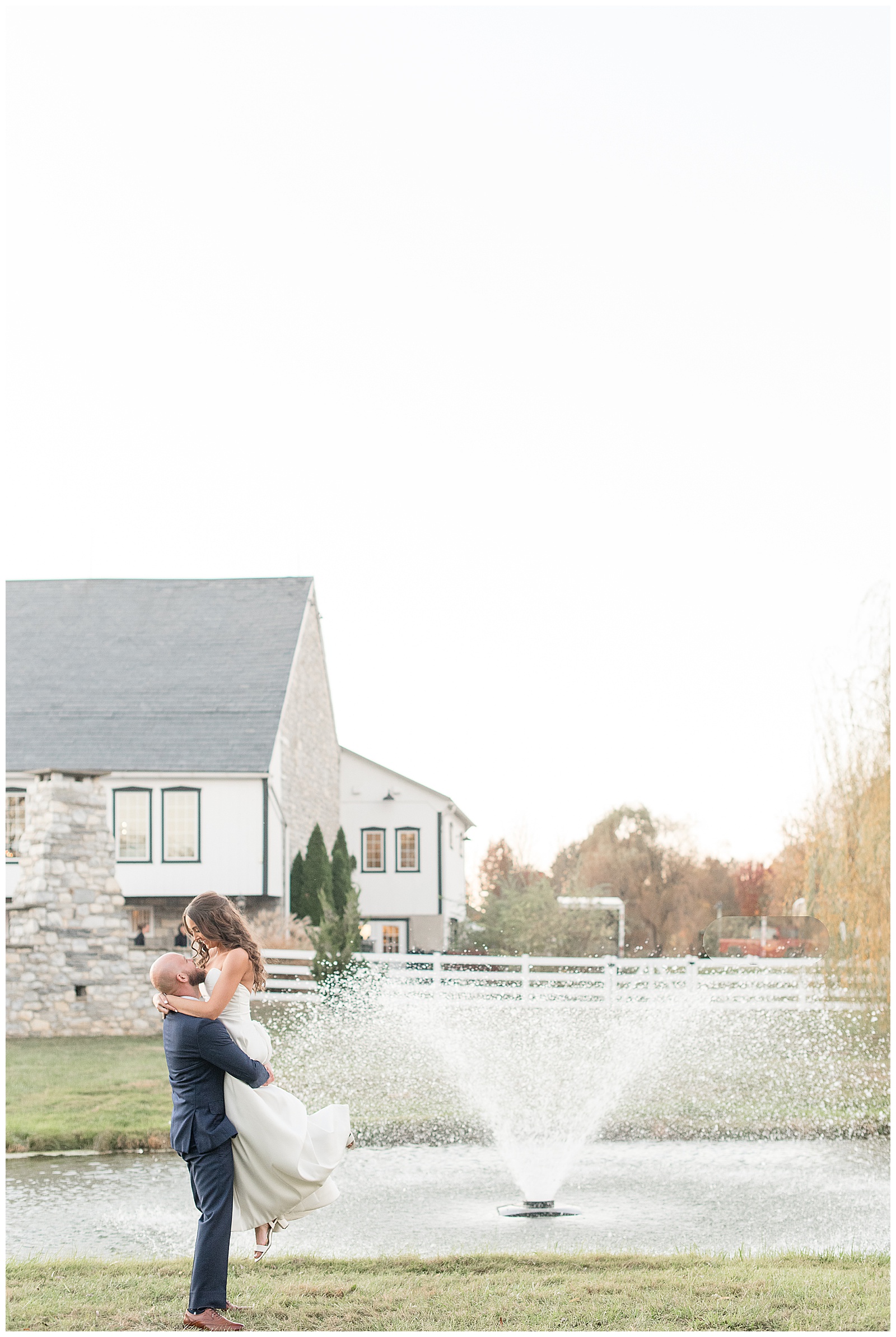 groom lifting his bride by pond with white barn in background at the barn at silverstone