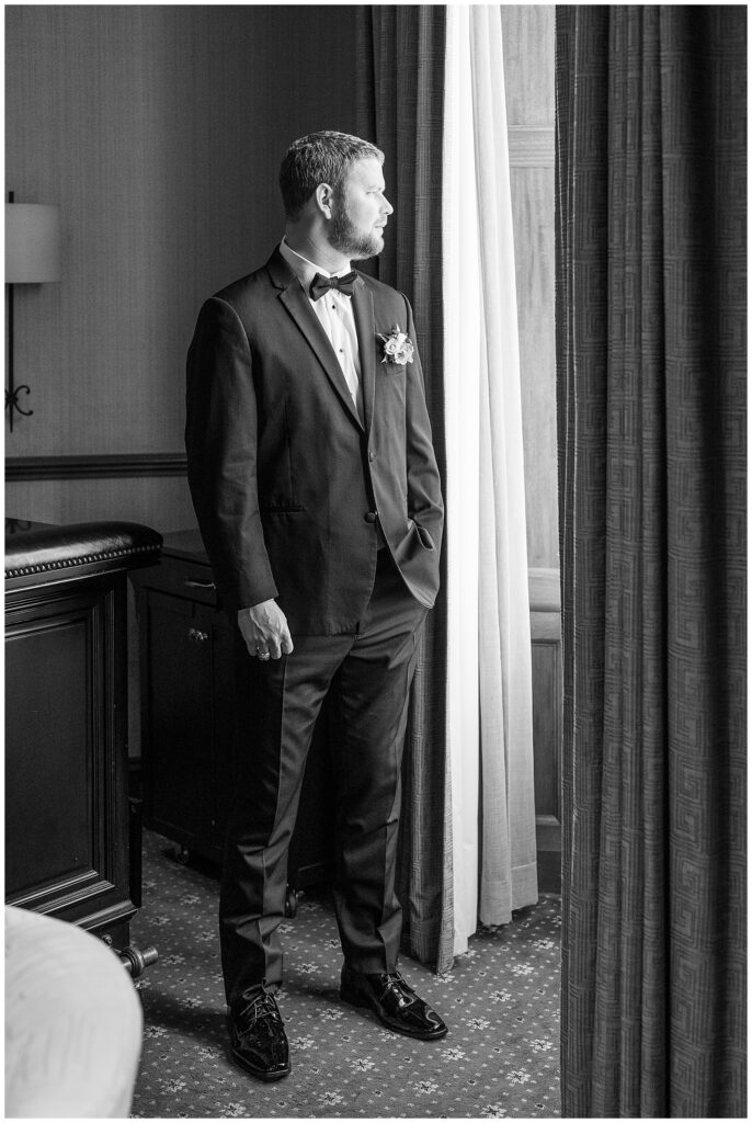 black and white photo of groom in his black suit looking left and staring out window at the lancaster marriott