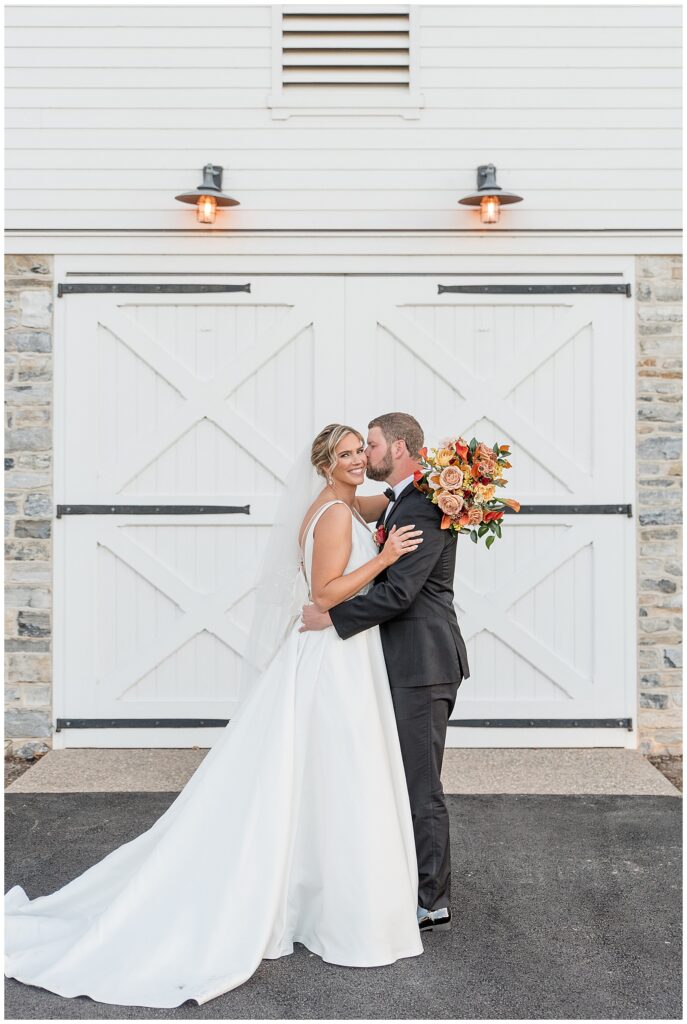 groom kisses his bride's left cheek by white barn doors at the star barn in lancaster pa