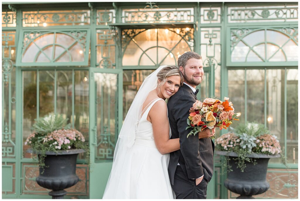 bride hugging her groom from behind as they both smile by glass building at the star barn venue in lancaster pa