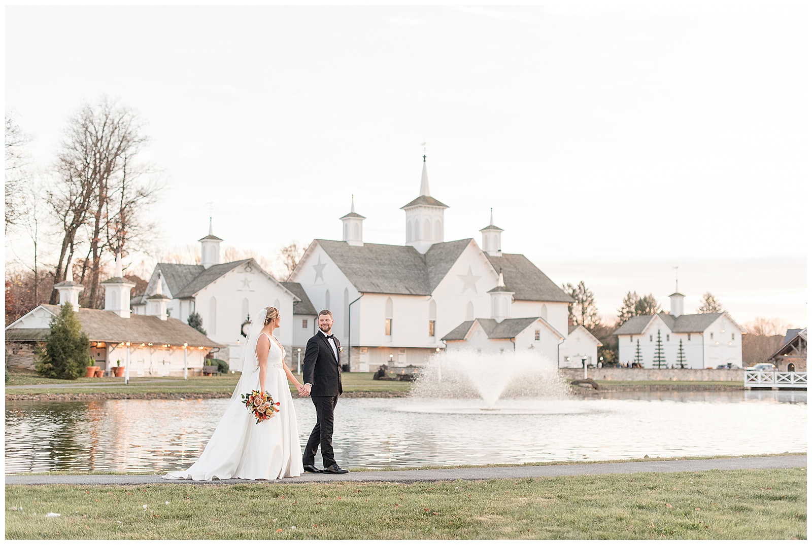 couple holding hands and walking along edge of pond wit star barn behind them in lancaster pennsylvania