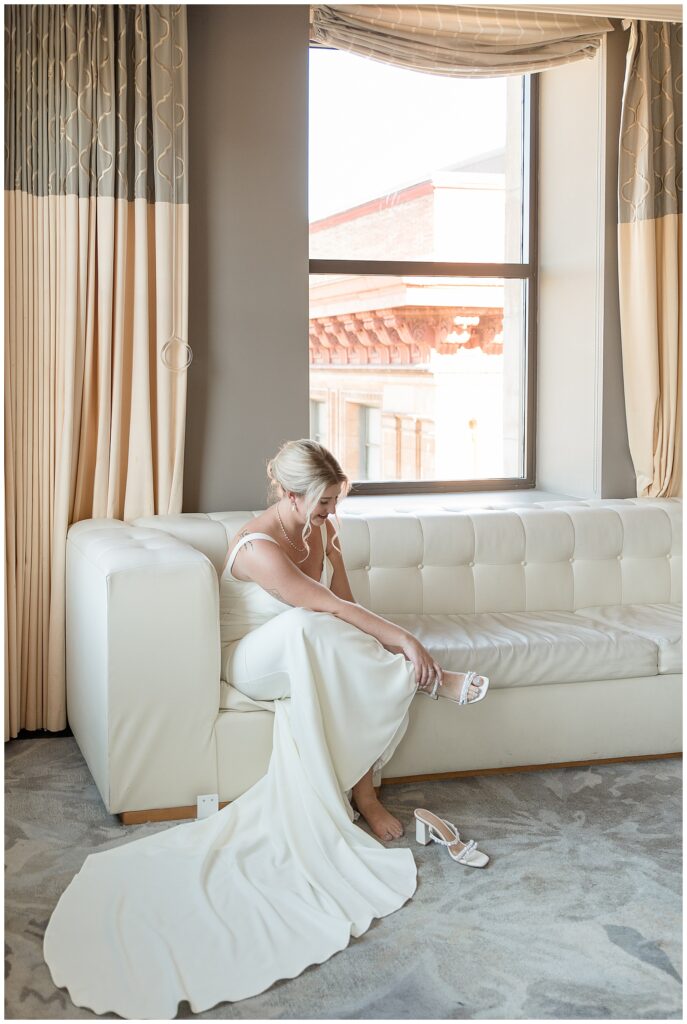 bride in white gown with spaghetti straps sitting on trendy white couch and putting white shoe on her right foot at kimpton hotel monaco