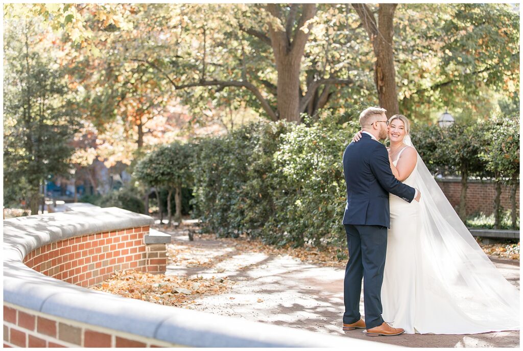 groom hugs his bride as she smiles as they stand on brick walkway by trees at hotel monaco in philly