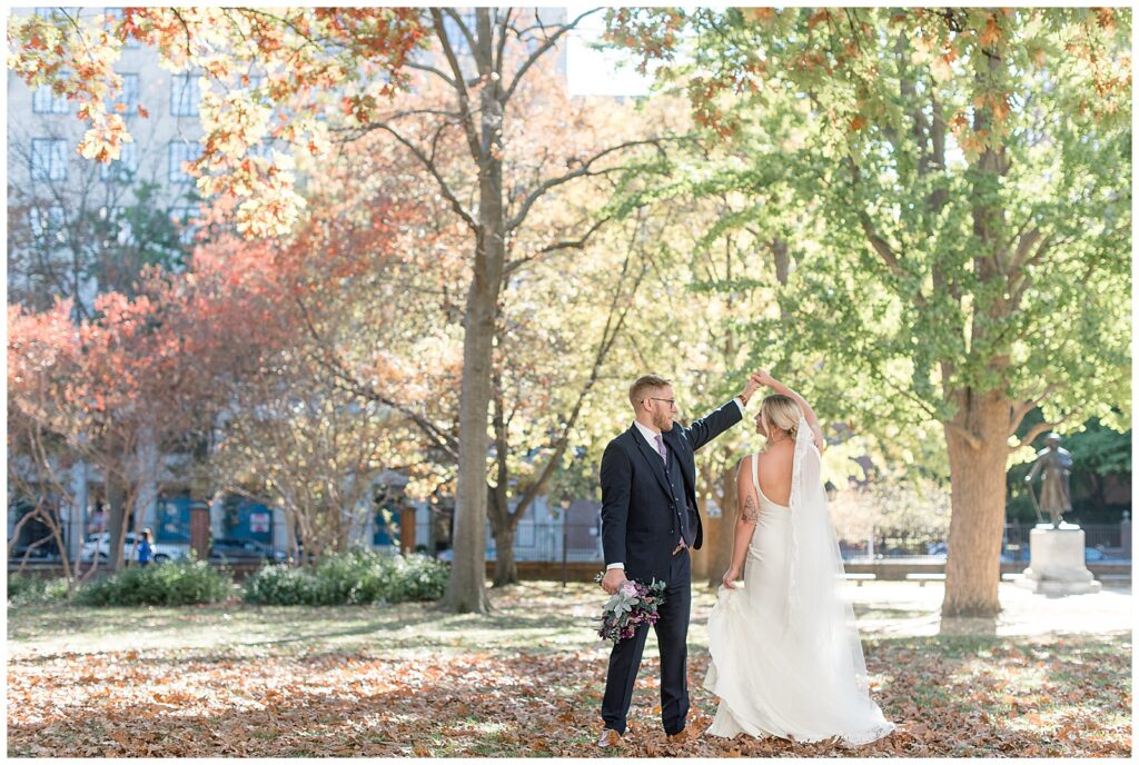 groom twirls his bride under his left hand by colorful fall trees in philly