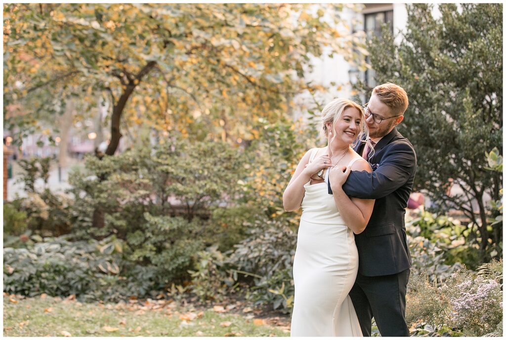 bride leaning back against her groom as he wraps his arms around her by colorful fall trees in philly