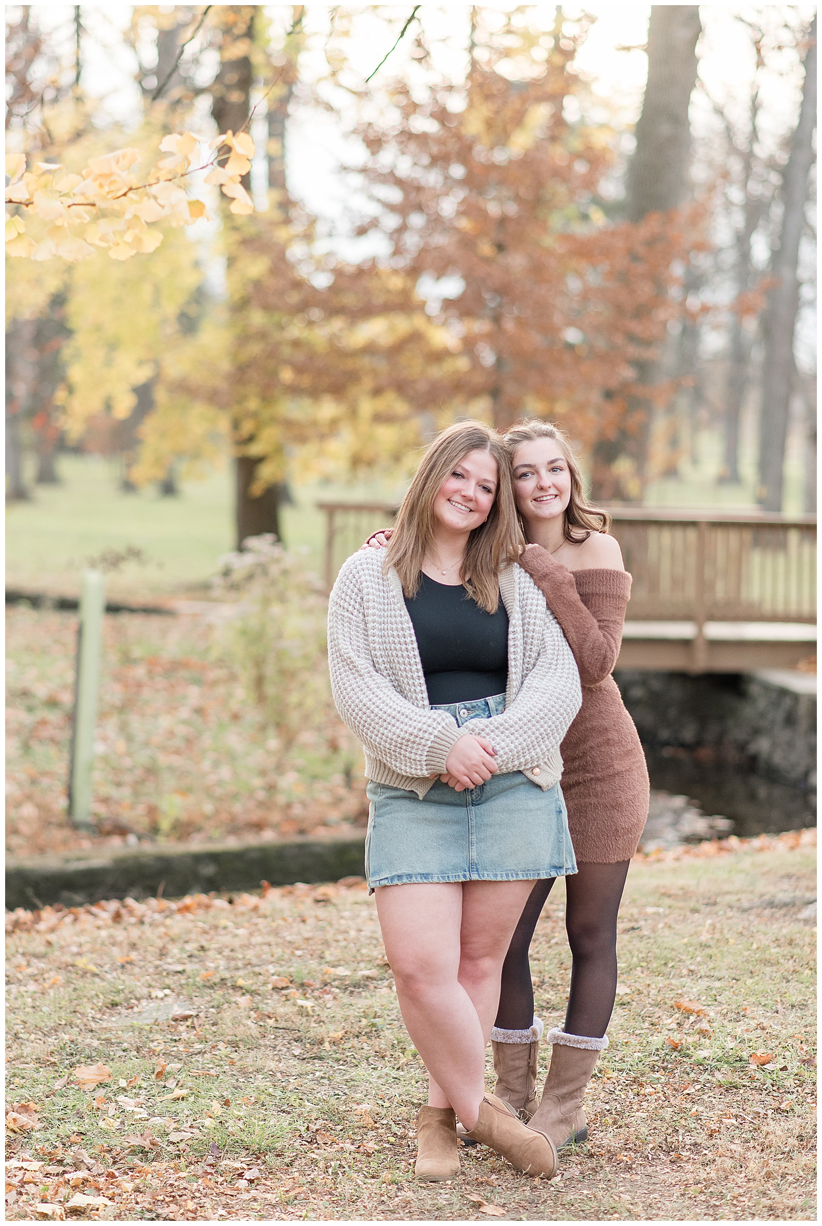 two senior girls leaning against each other with colorful fall trees behind them at longs park