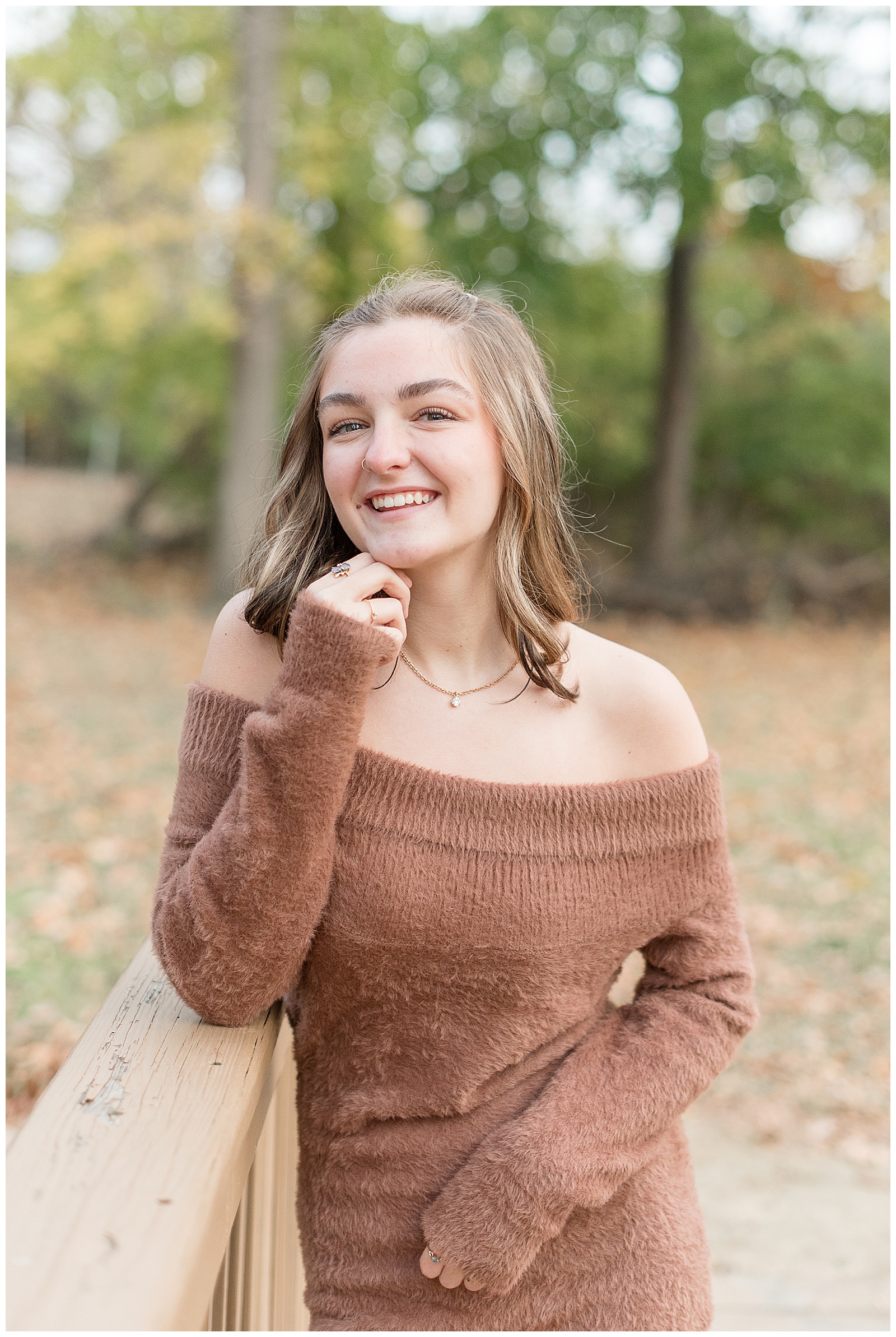 senior girl wearing off the shoulder brown sweater leaning against wooden bridge at long's park