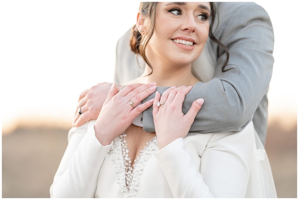 closeup photo of groom hugging his bride from behind as she holds his left arm and looks to the left in field in lancaster pa