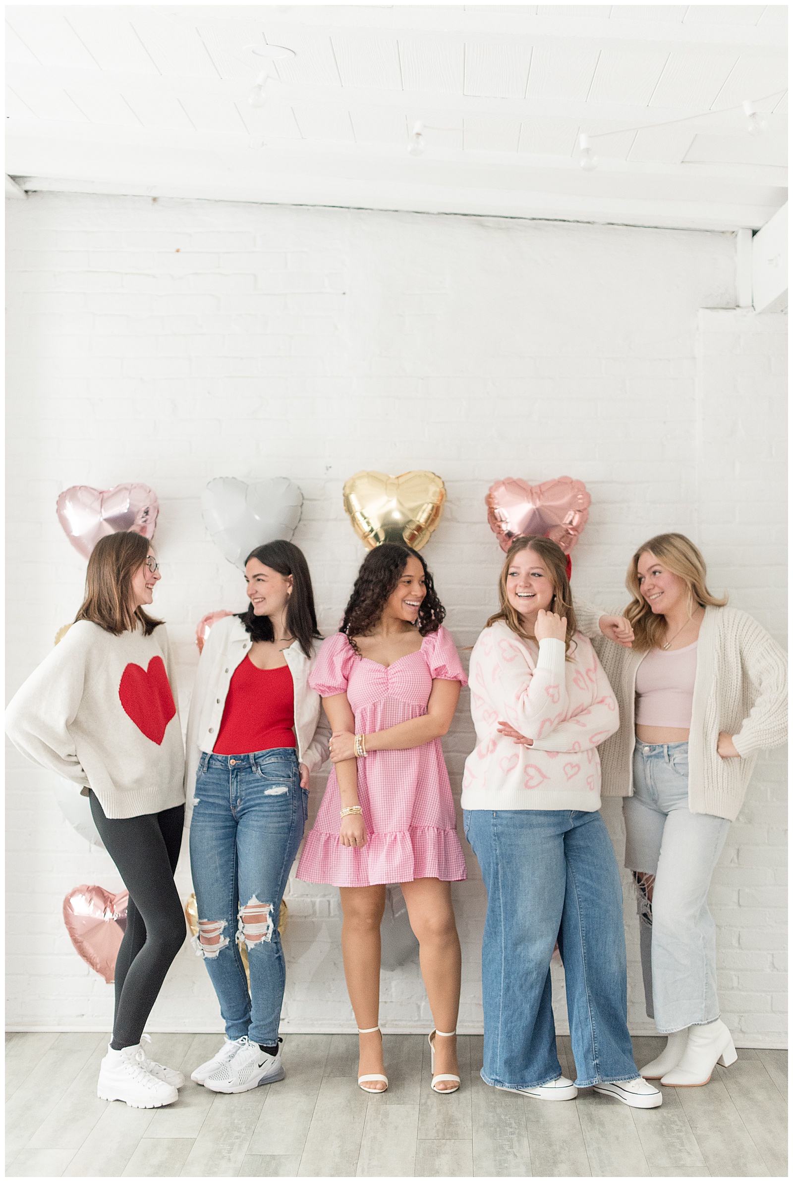 five female seniors standing by white wall with metallic helium balloons on wall behind them as they look at each other at the white room