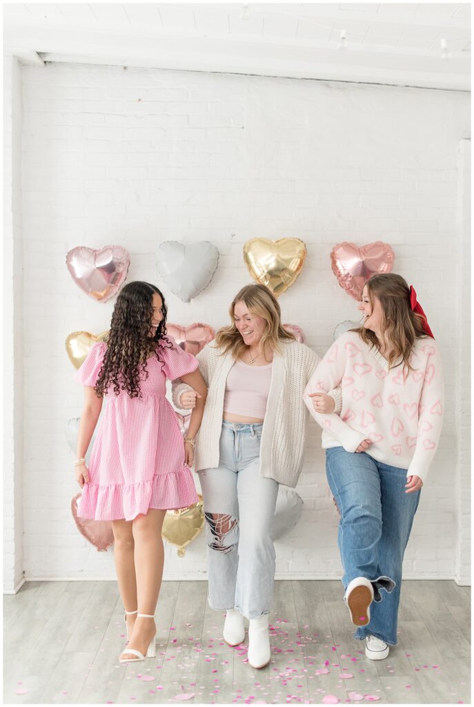 three senior spokesmodels with arms linked as they smile and look at each other by heart balloon wall at the white room