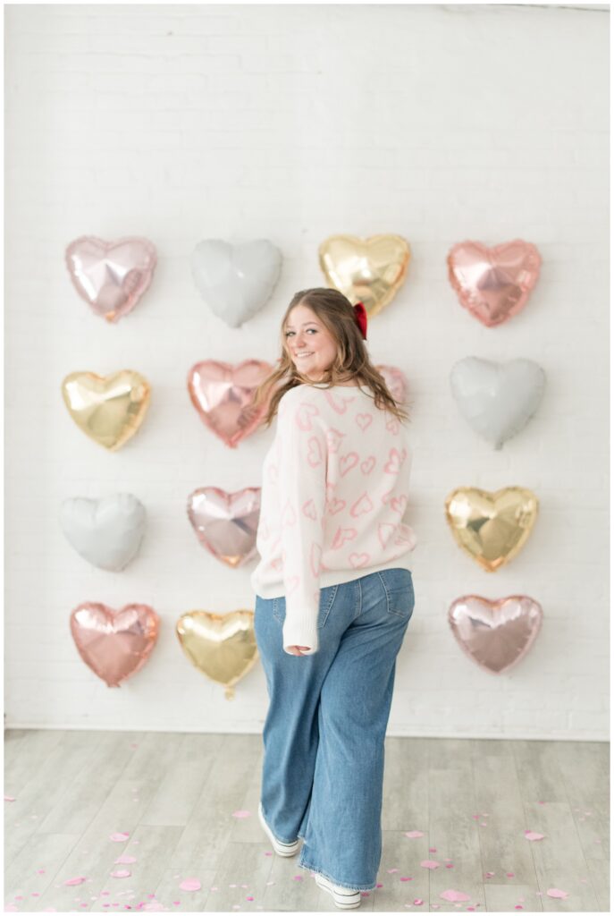 senior girl with back toward camera looking back over left shoulder by heart shaped balloon-covered wall in lancaster city