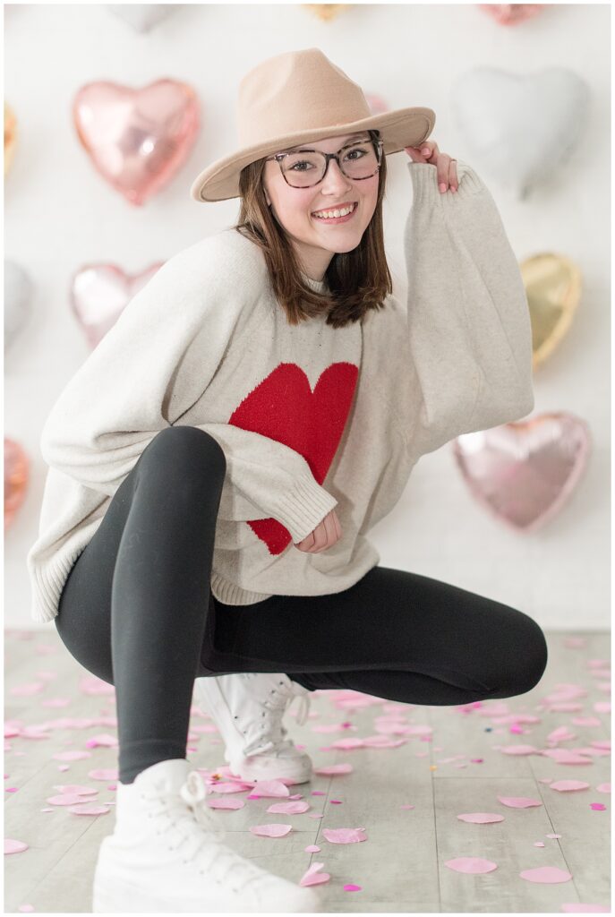 senior girl crouching down with right foot forward wearing heart sweater and holding cute hat with her left hand at valentine's day shoot in the white room