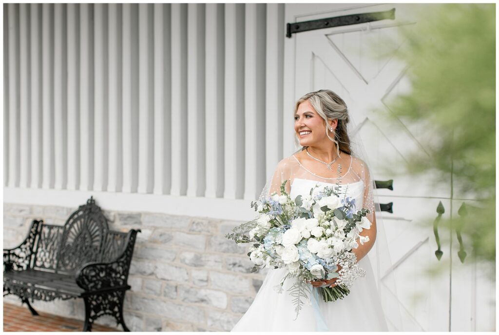 bride in beautiful white gown with large bouquet of blue and white flowers by white barn in lancaster pennsylvania