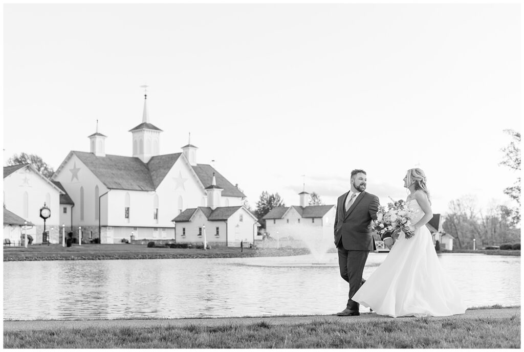 black and white photo of couple holding hands and walking along pond with the star barn in the background