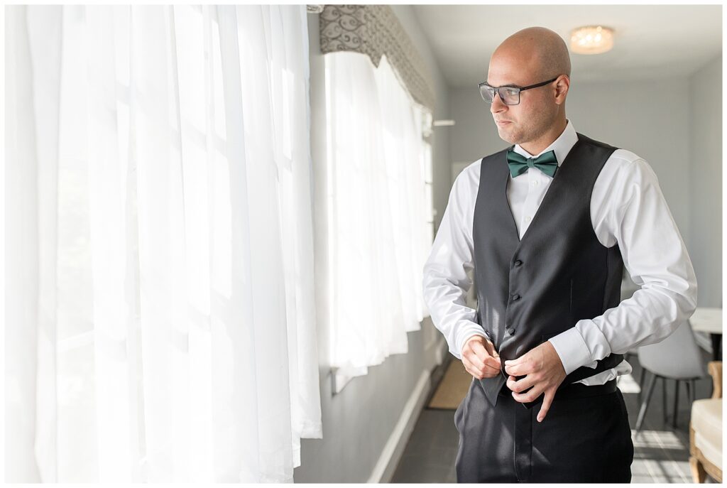 groom buttoning up his black vest by window at lauxmont farms in york county pennsylvania